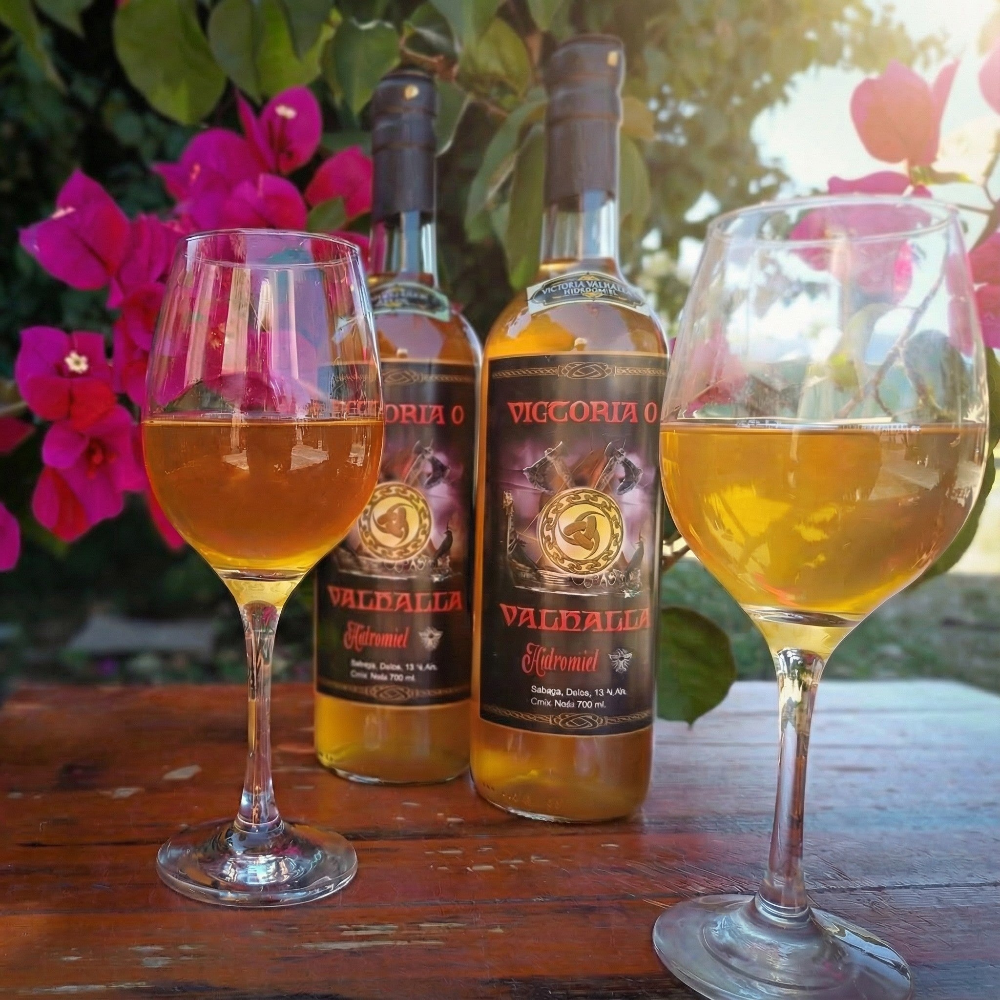 Dos copas de hidromiel y Dos botellas de Hidromiel Victoria o Valhalla sobre mesa rustica con un fondo de flores santaritas rojas. Imagen totalmente natural.