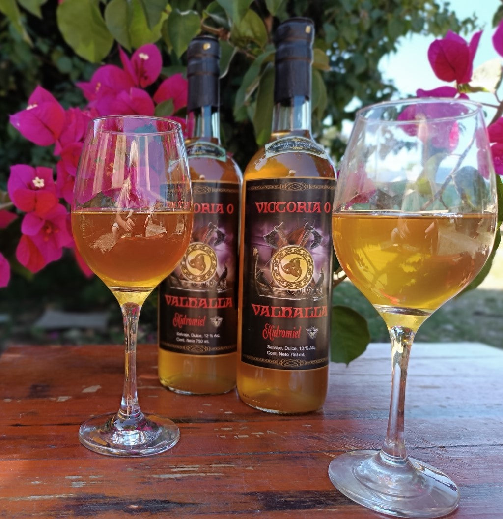 Dos copas de hidromiel y Dos botellas de Hidromiel Victoria o Valhalla sobre mesa rustica con un fondo de flores santas ritas rojas. Imagen totalmente natural.